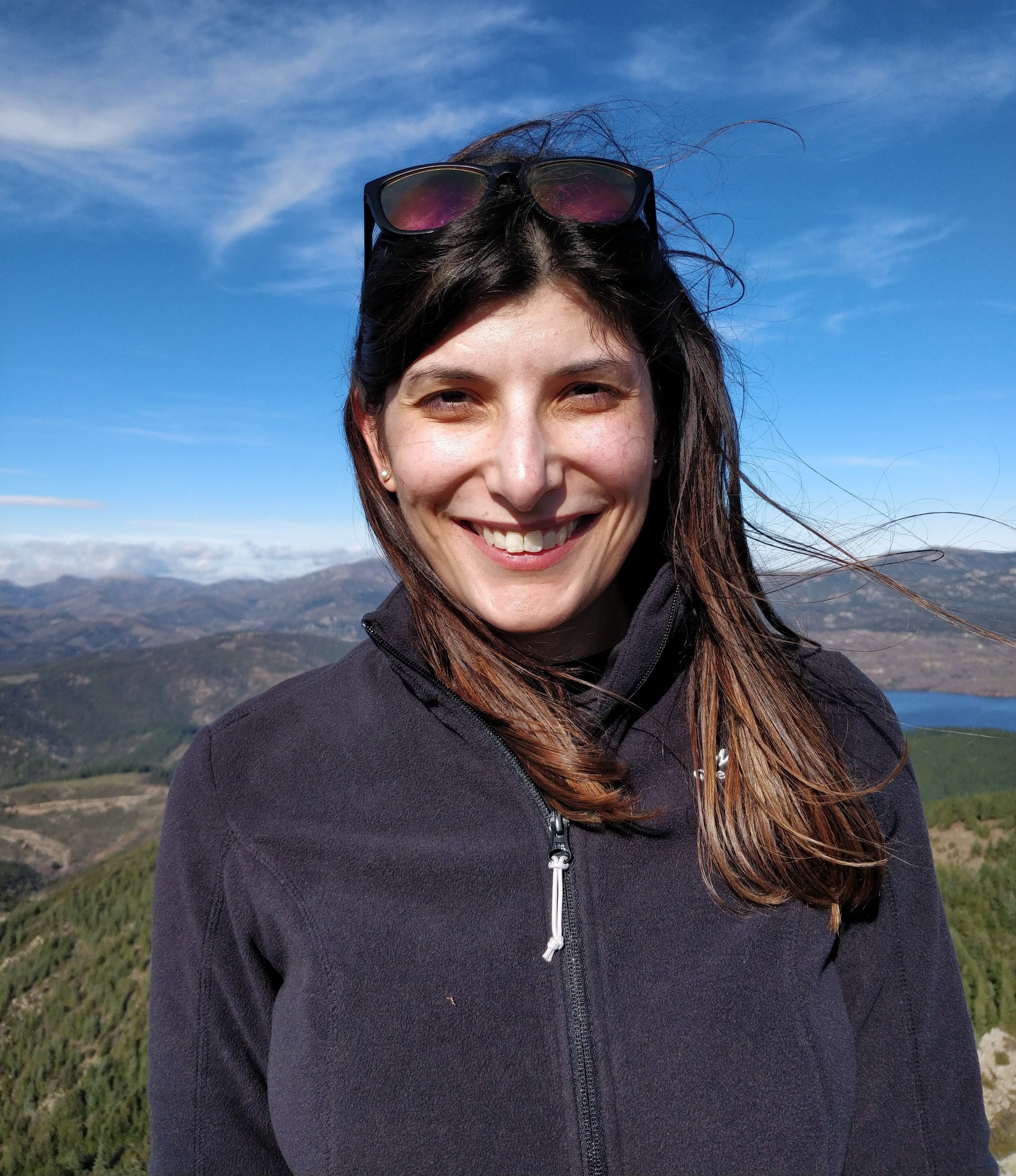 Image of woman standing outside with sunglasses on her head, blue sky and hills in the background