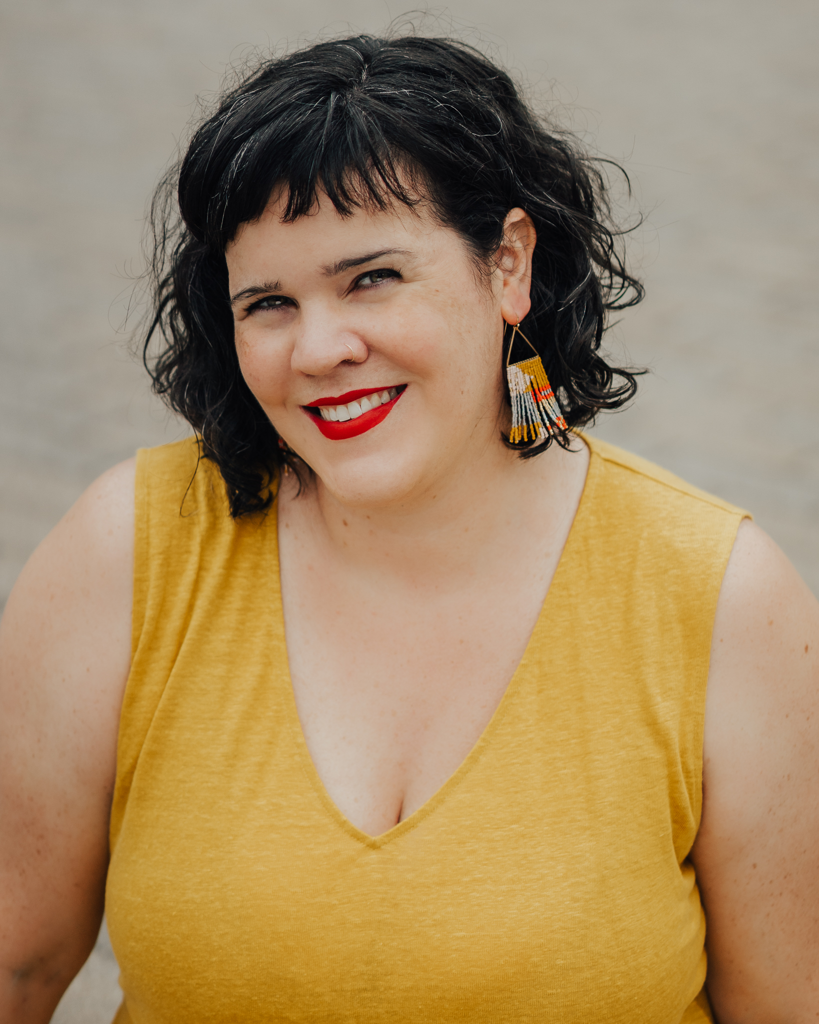 A picture of Letitia Henville, a white lady with black-and-grey hair who is wearing a gold nose ring, bright red lipstick, and a yellow sleeveless shirt. 