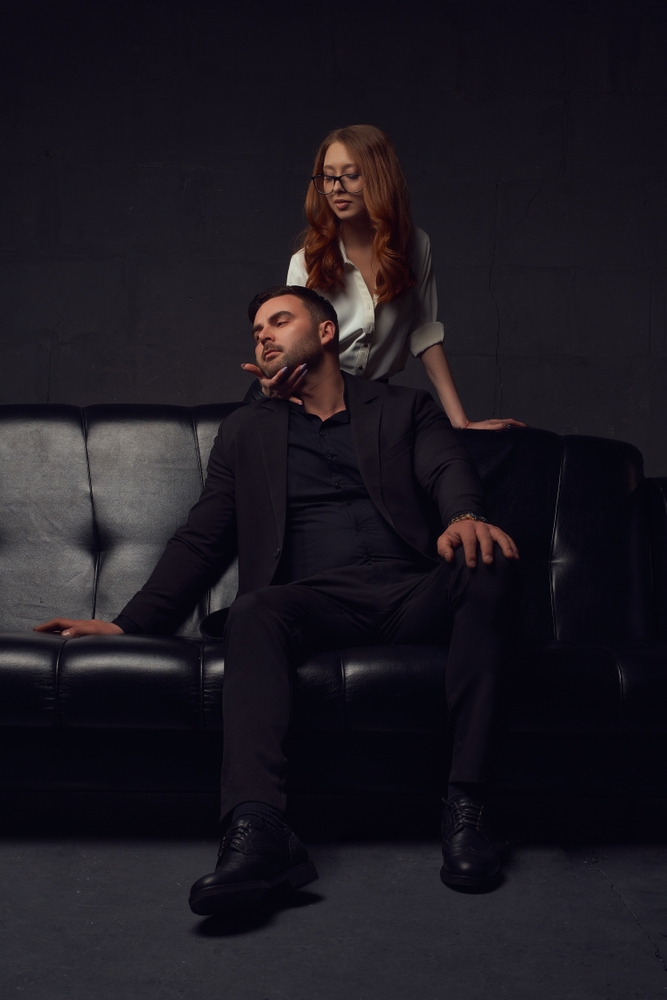 A white woman with long red hair and glasses, takes a Latino man with a light facial hair and a dark suit by the chin as he sits on a black leather couch. 