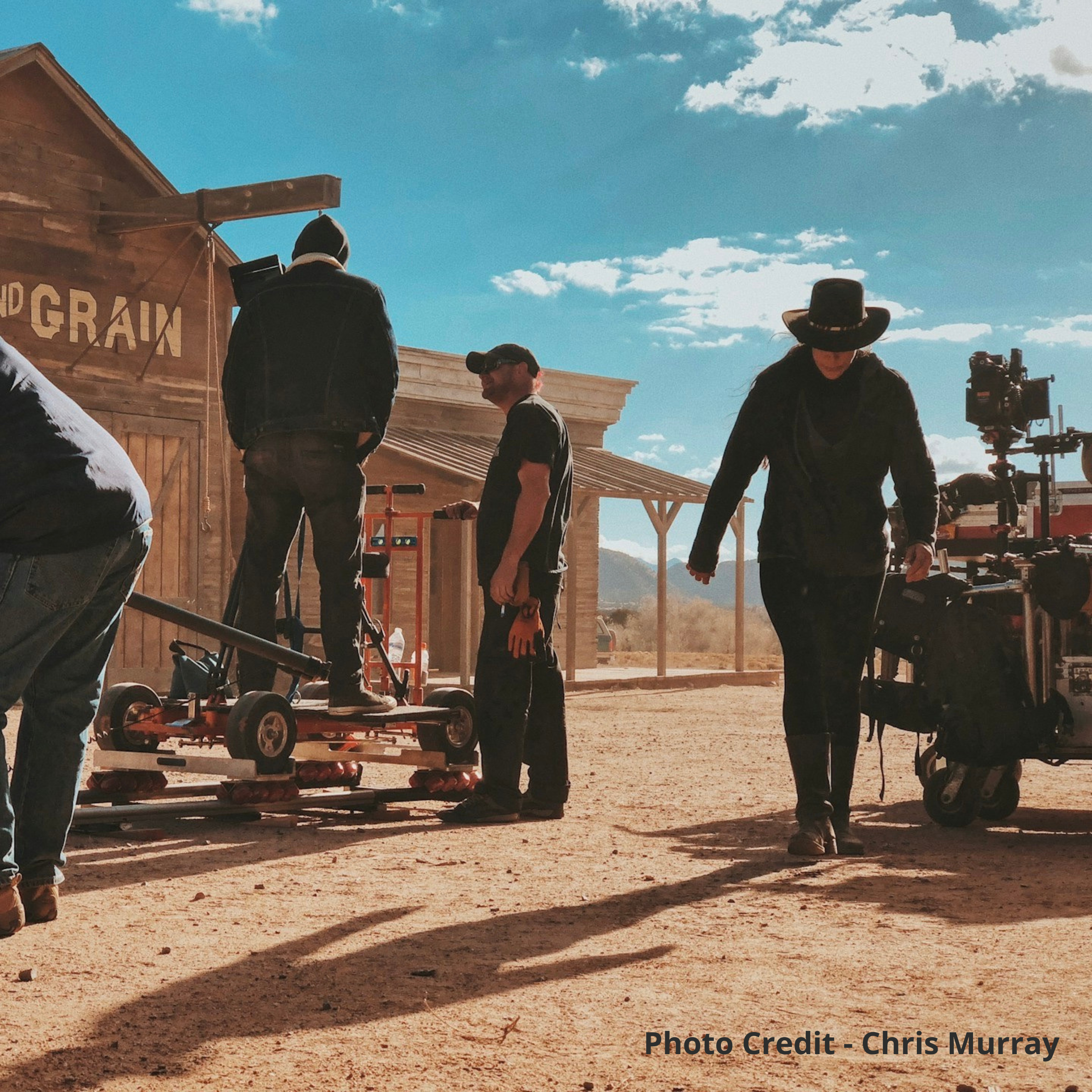 Three people in black standing in front of a old grain store movie set in the desert with a camera dolly and cart holding camera equipment