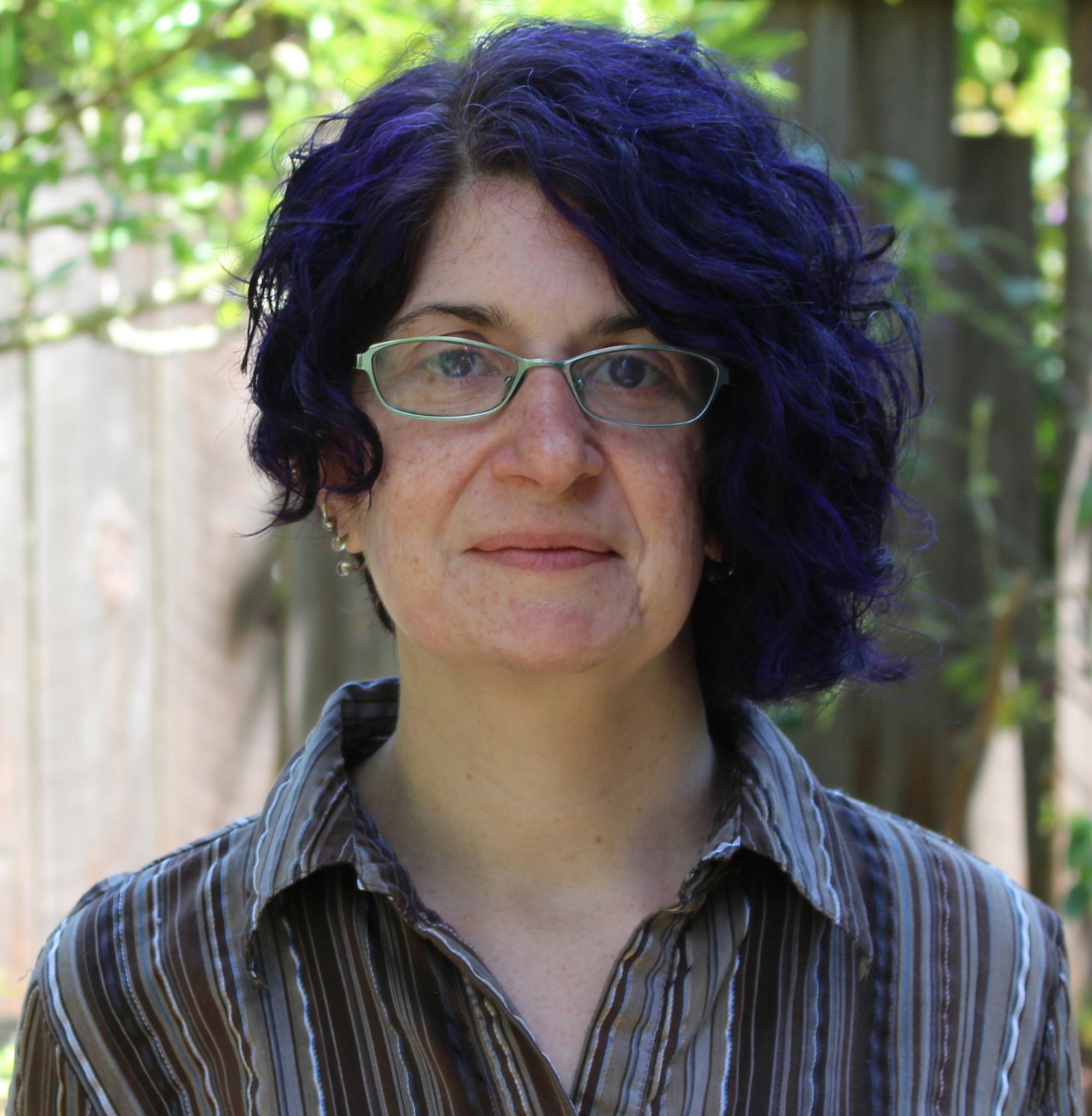 Rachel, a White woman with curly purple hair and glasses, in front of a tree and fence, wearing a warm gaze