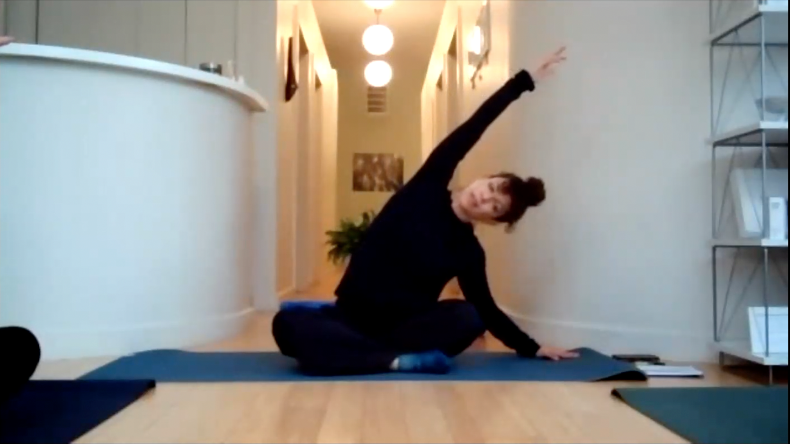 Teacher sitting on a yoga mat stretching upper body to one side with her right arm above head.