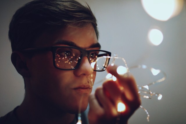 A person with glasses is holding small string lights close to their face. The lights are reflecting off the glasses.