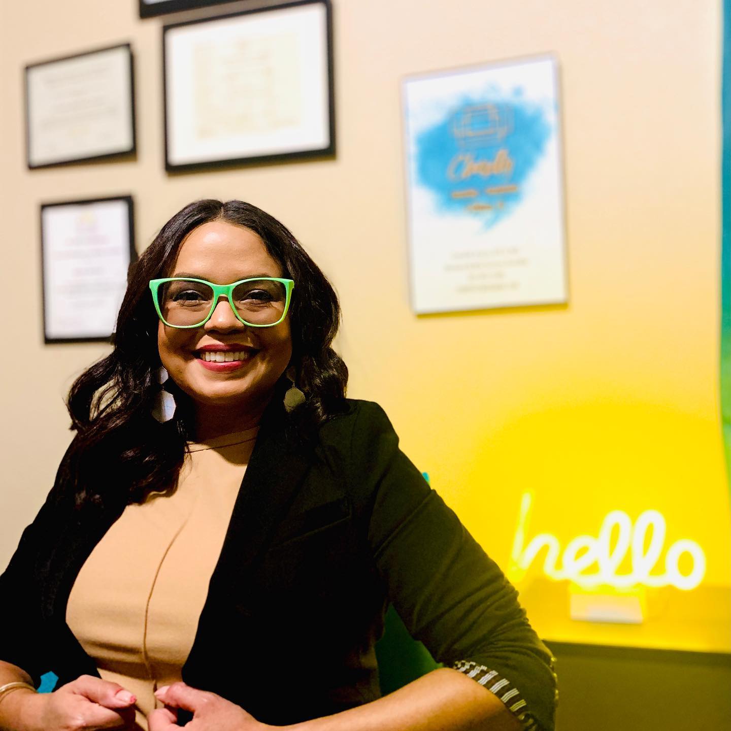 Latina Woman with green glasses sitting on a chair. She is sitting next to a yellow neon sign that says &quot;hello&quot;.