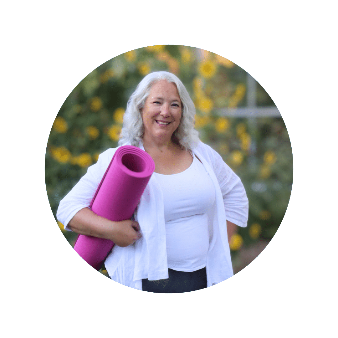 Aruna stands in front of yellow daisies holding a pink yoga mat under her arm and smiling