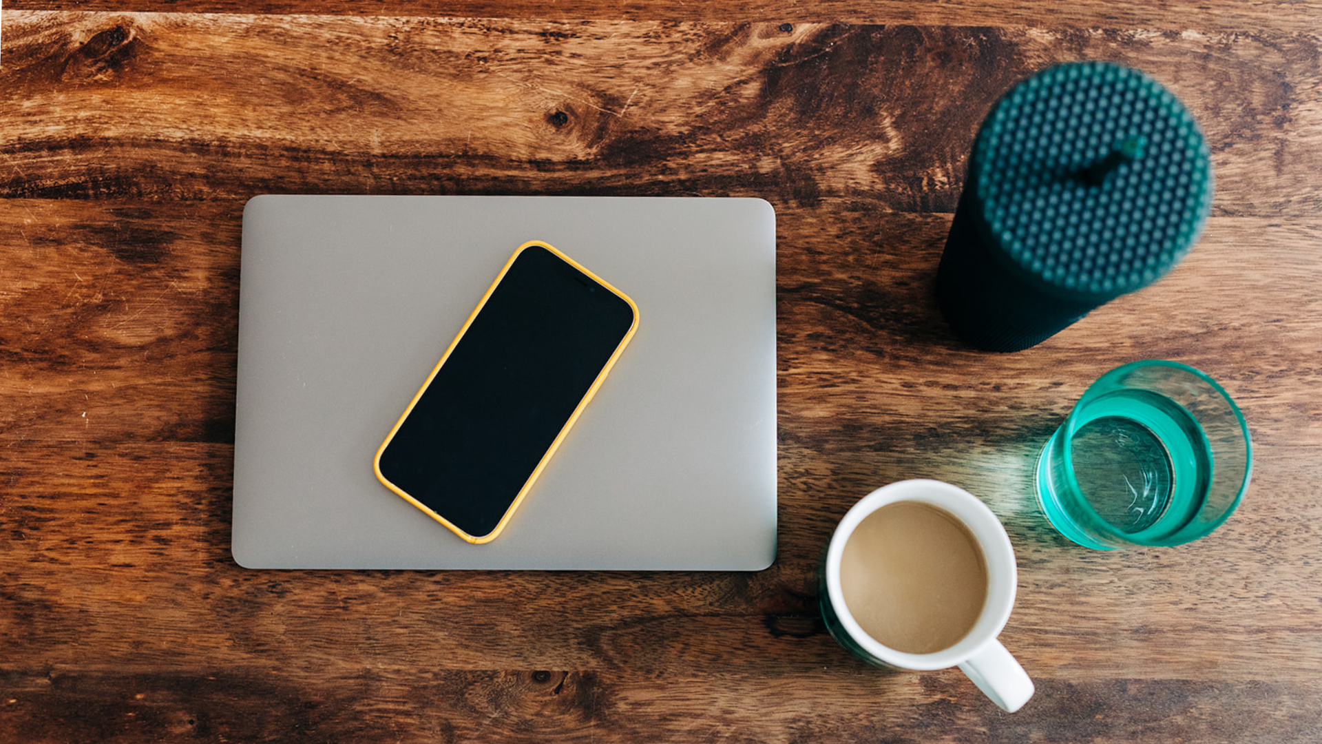 Birds eye view of a wood desk with a macbook, a green glass filled with water, a white mug filled with coffee and a textured dark green water cup with a lid and a straw.