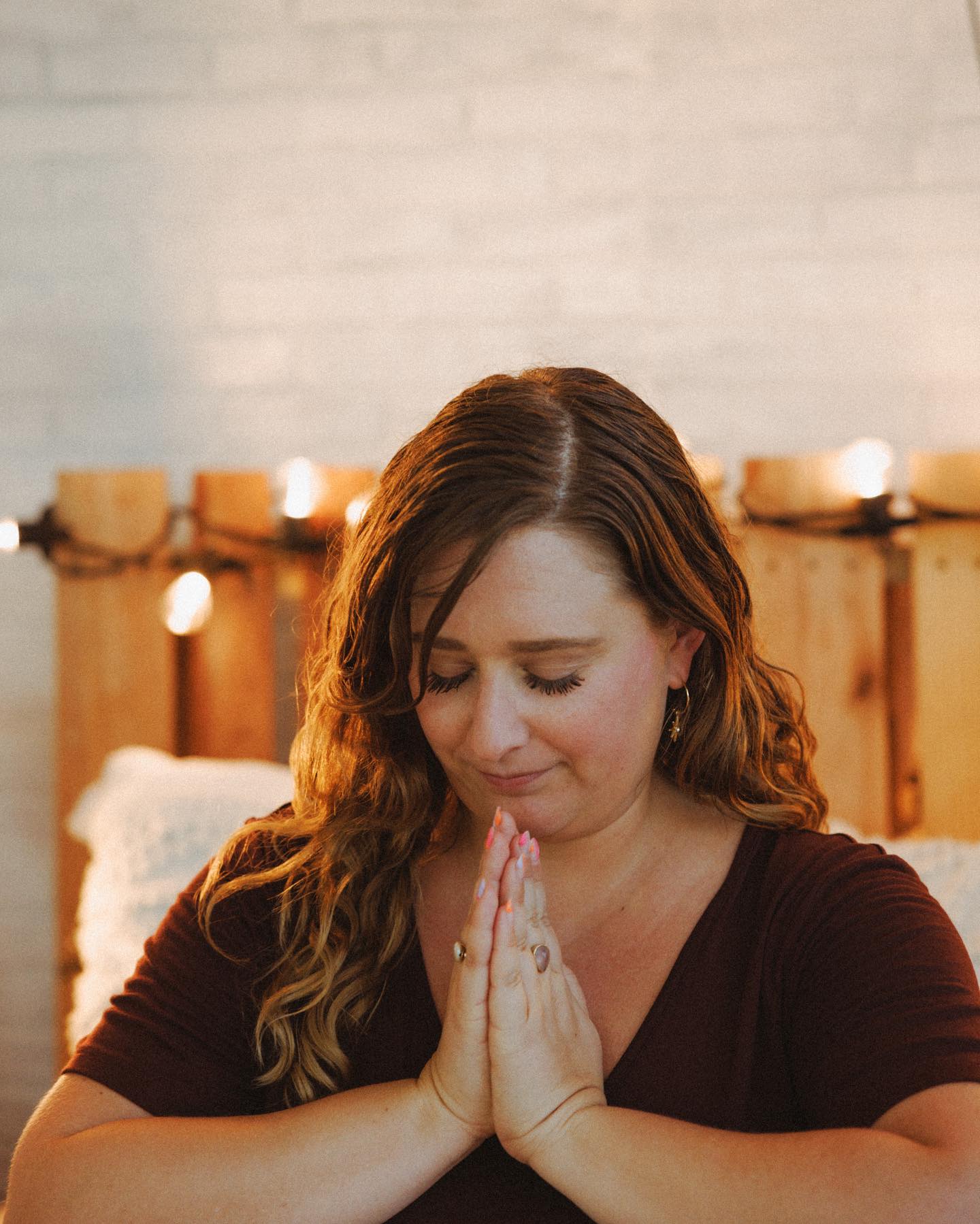 A dirty blonde haired white lady is meditating behind a wooden bed frame with lighting. She is wearing a maroon v shirt with gold rings and earrings.