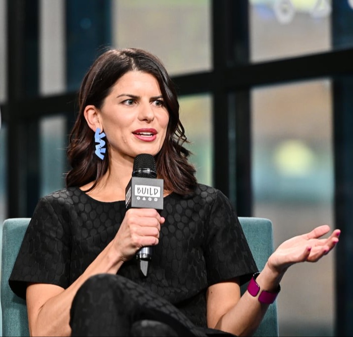 Margaux Joffe speaks into the microphone at Build Studios in New York. She wears a black textured suit and blue geometric earrings.