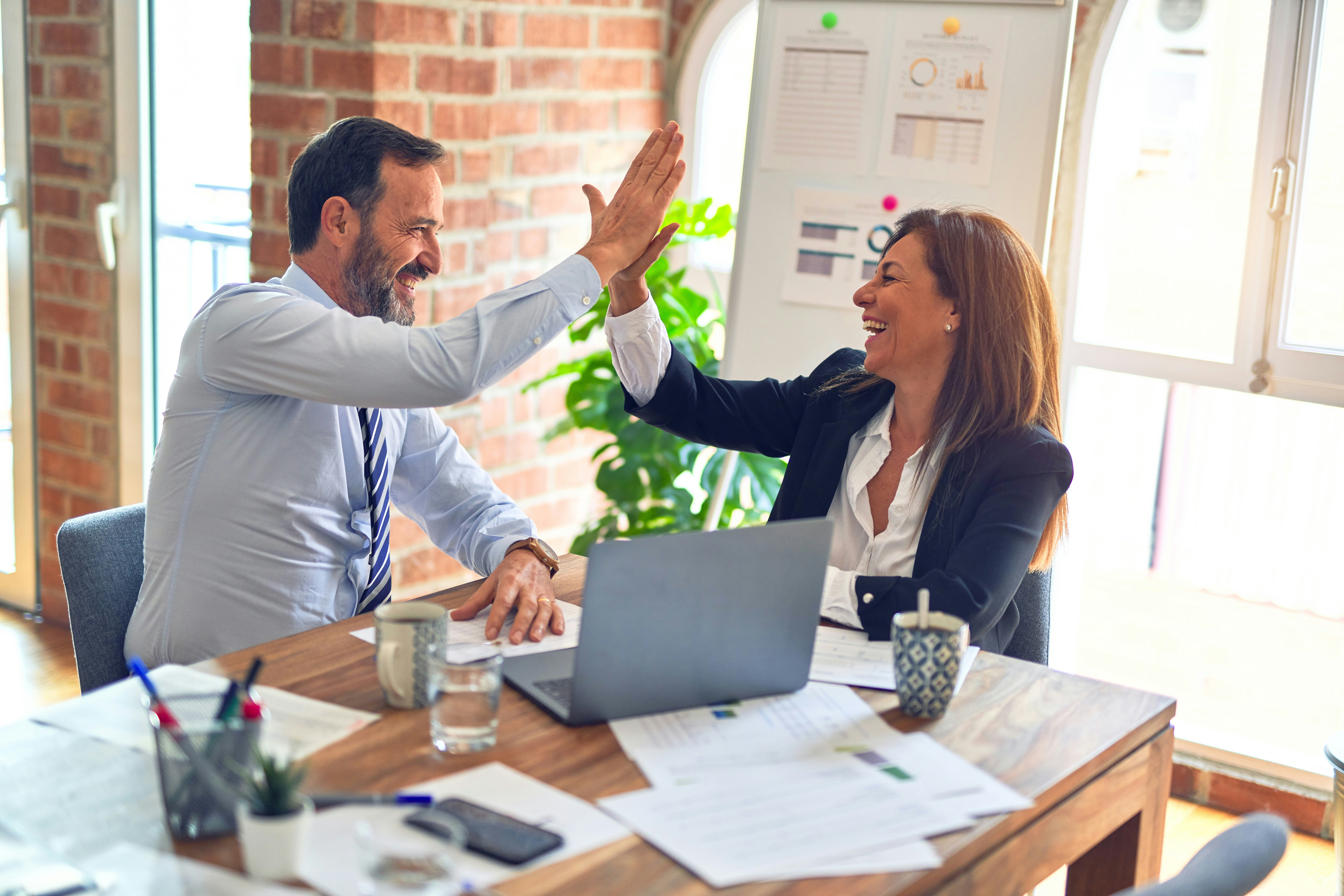 A man in a blue dress shirt and tie is high-fiving a woman in a white blouse and blue jacket. They are smiling behind a laptop. The background is a bright, modern office with some classic brick elements.