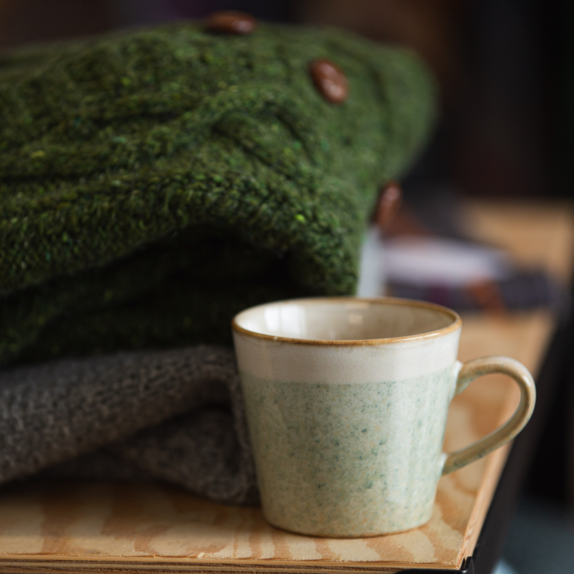 Coffee mug with folded pile of knitted garments behind it on a desk.