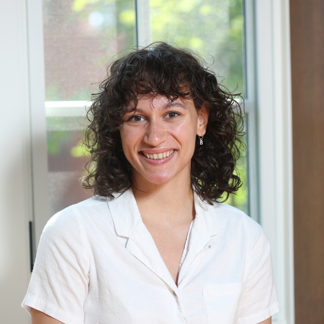 A woman with curly dark hair wearing a white shirt in front of a window with greenery outside.