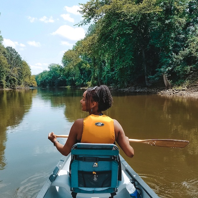 Black woman with micro twists holding a paddle in a canoe on a river, she is wearing a life jacket