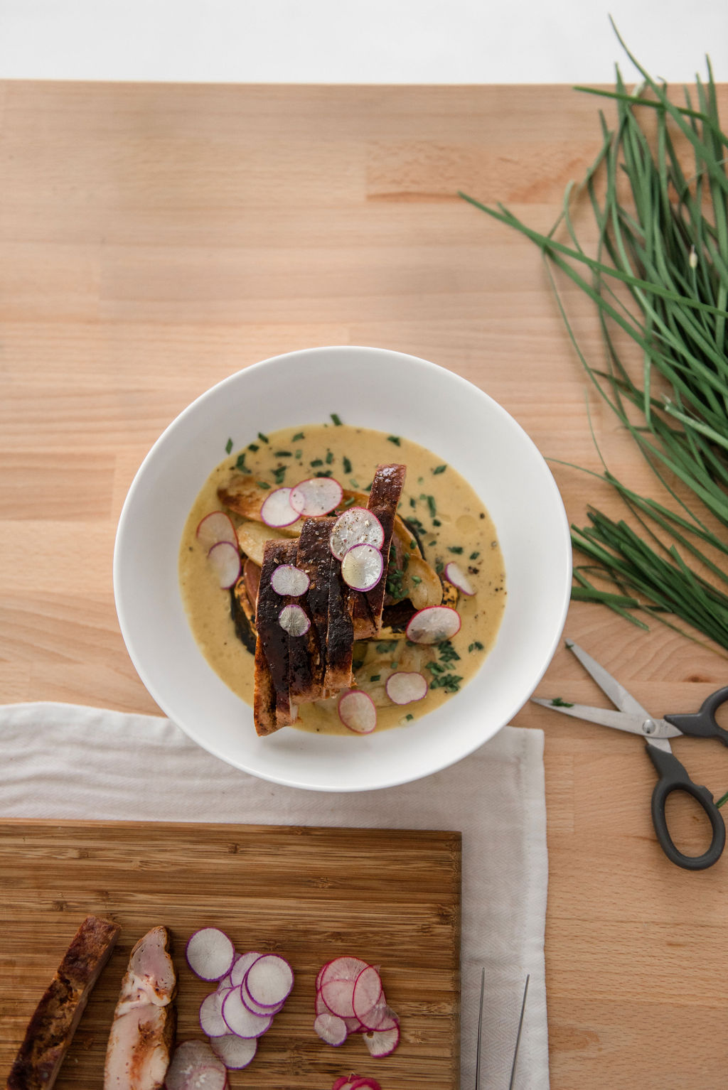 A serving bowl with seared meat, sauce and radish garnish sits on a butcher block table with herbs, scissors, a cutting board and other ingredients.