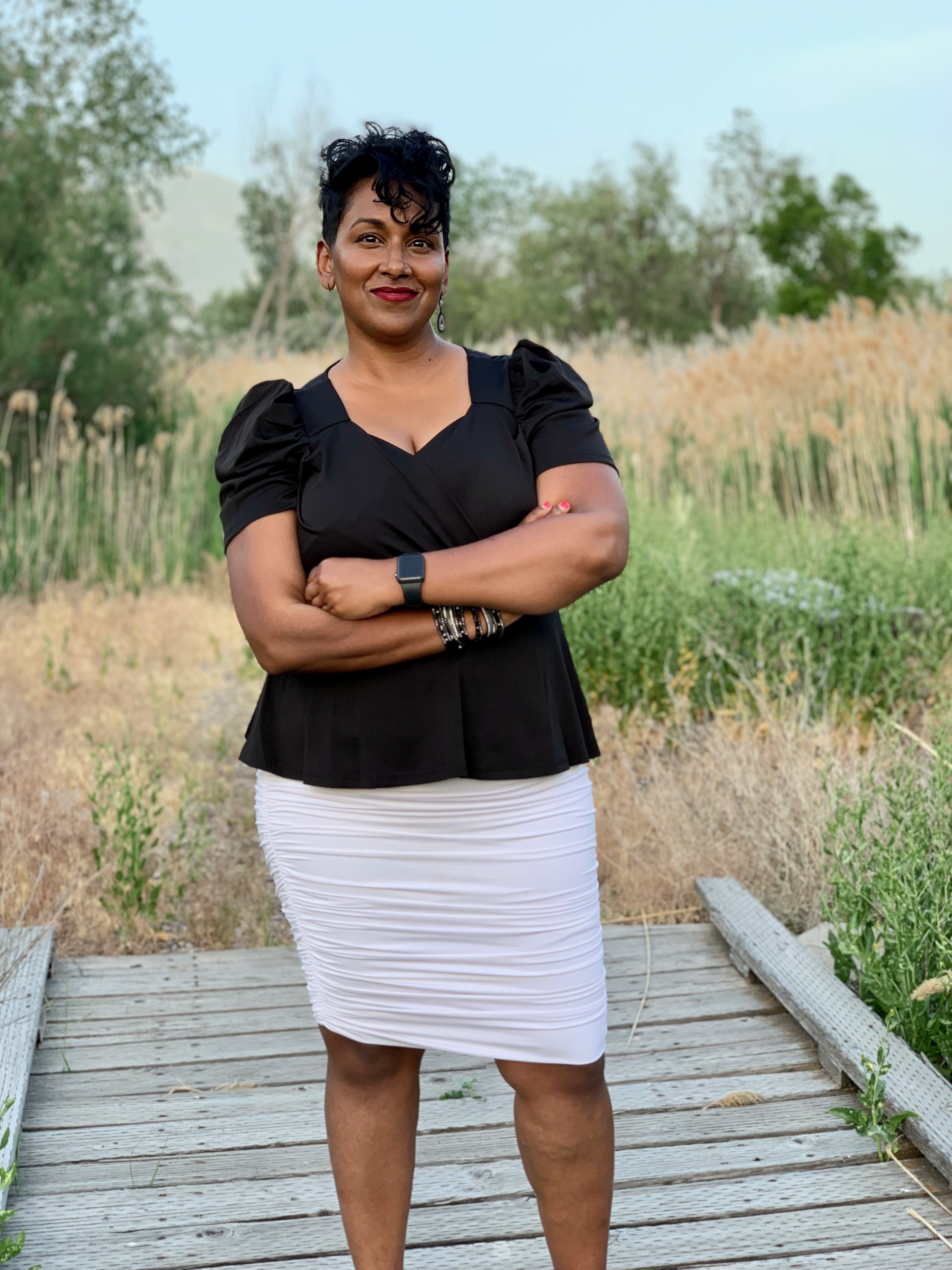 photo of Leatha, a woman of South Asian descent, with brown skin and short black curly hair, folding her arms and looking at the camera. She is wearing a black shirt and white skirt, and she is standing in a field.