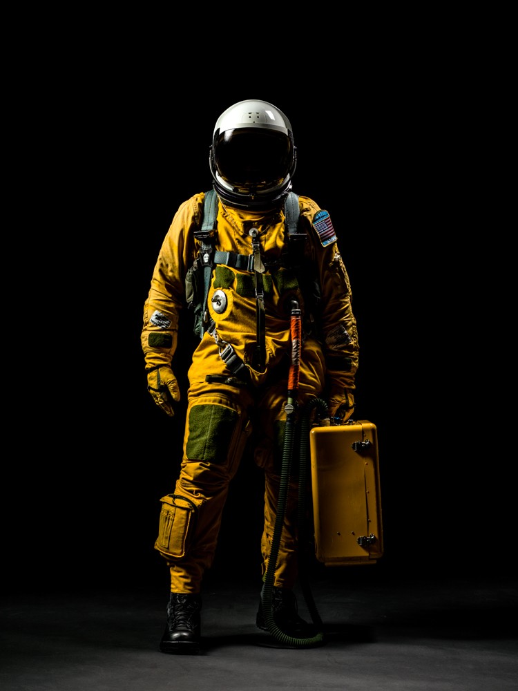 Full figure standing in orange spacesuit with a life support unit in the left hand and black background.