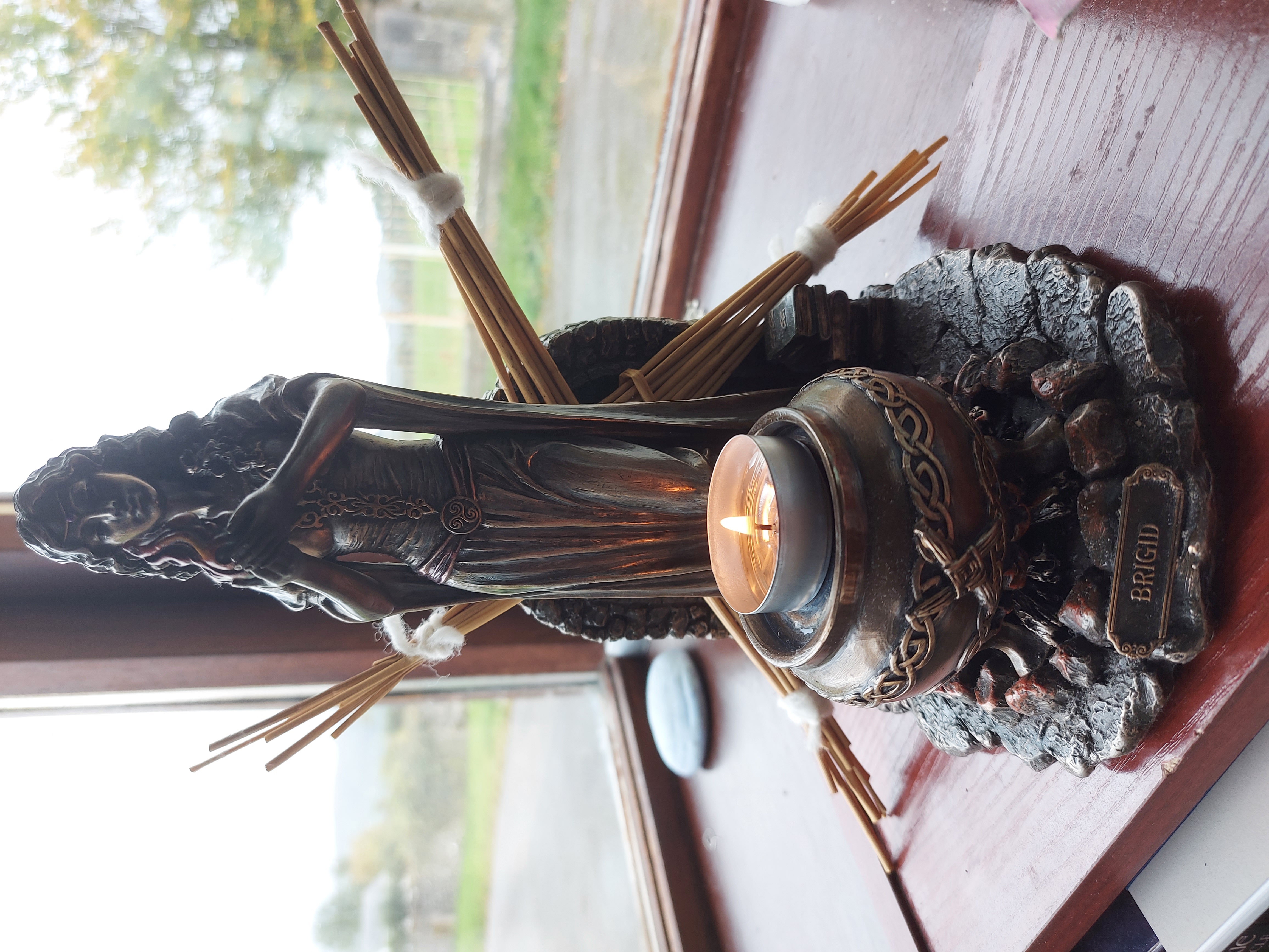 Photo shows an image of my bronze Brigid statue. She is holding a flame in her hands, wearing a long flowing dress with a belt or girdle around the waist and very long floor sweeping sleeves. She has long curly hair and a cauldron is in front of her, with a candle burning on the top of the cauldron. There is a pile of books (part of the statue) by her side. Propped up behind the figure, there is a 4-pronged Brigid&#39;s cross made from rushes and tied with white wool. At the back of the statue, there is a stone fireplace depicted, with flames within. 