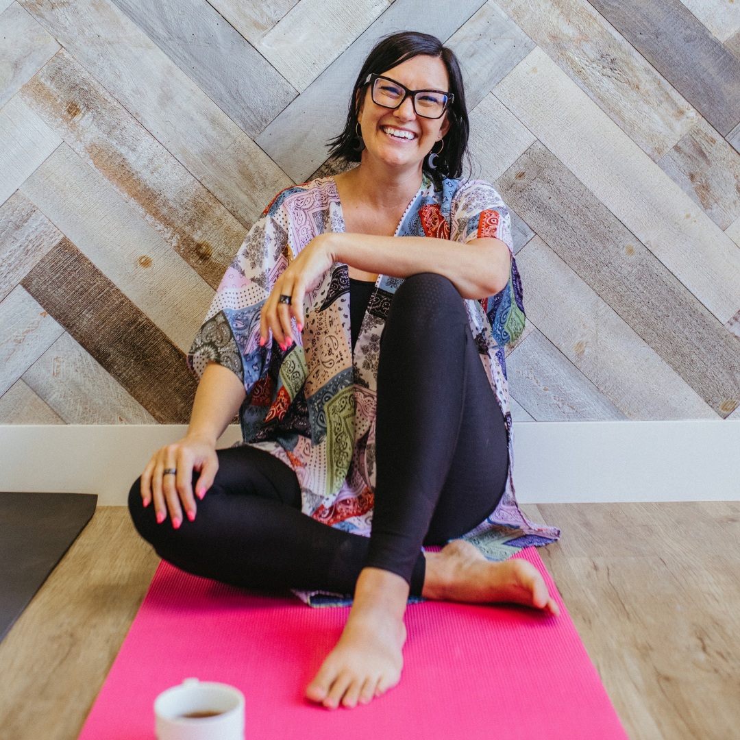 Melissa smiling at the camera with her arms folded in front of her. She is sitting on the floor, on a mat, wtih a cup of tea in front of her.