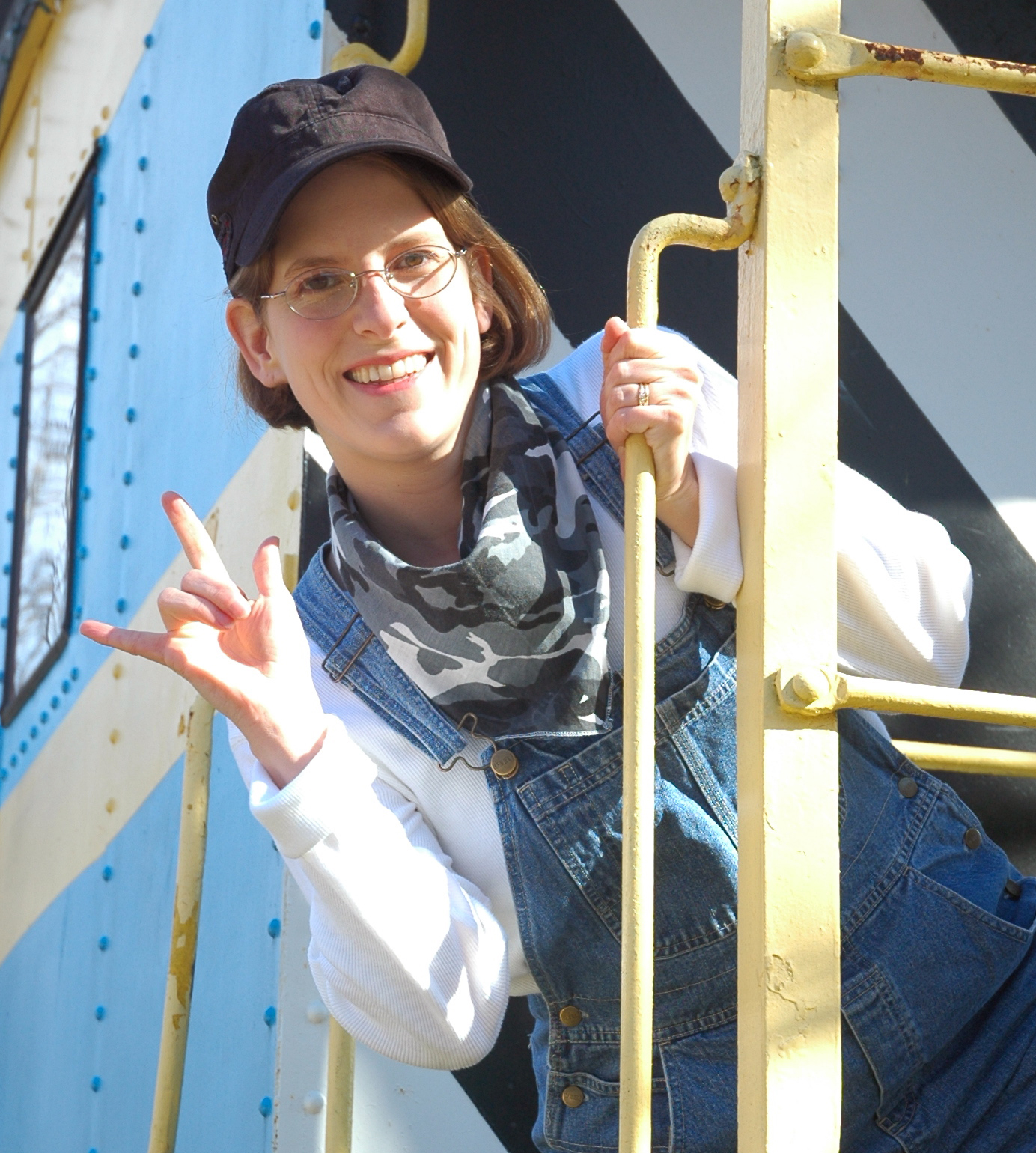 a white women with glasses and short brown hair leans off the back of a train caboose, signing I-LOVE-YOU in American Sign Language. She is wear a conductor&#39;s hat and overalls.