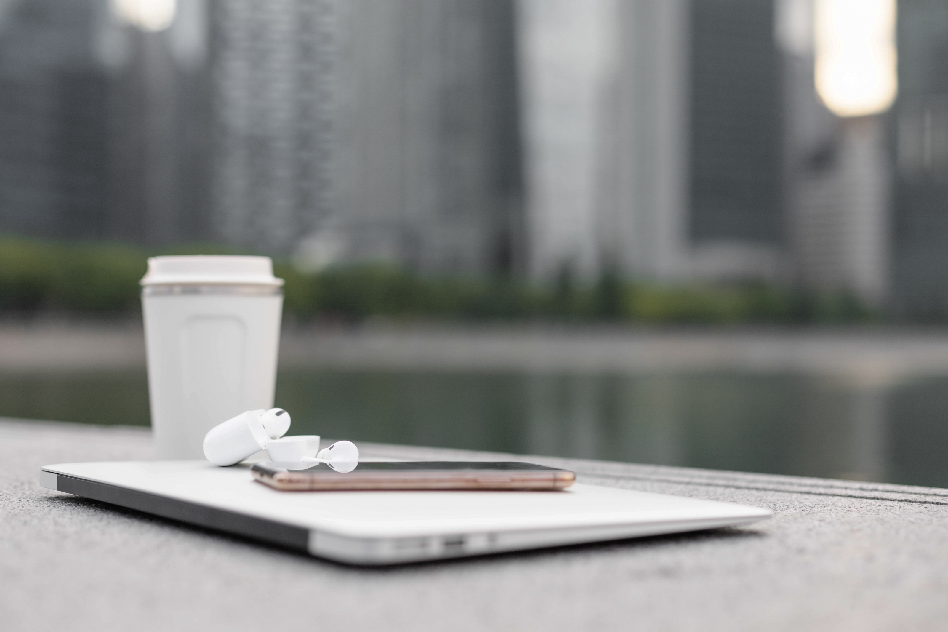 An image of a reusable cup near a closed laptop on a table. A mobile phone and wireless headphones are on top of the computer. The background is a blurred outdoor scene.