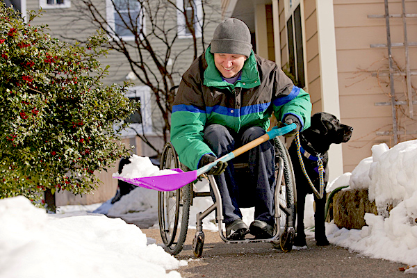 Person in a wheelchair using a shovel to clear a path snow, with a black dog sitting alongside