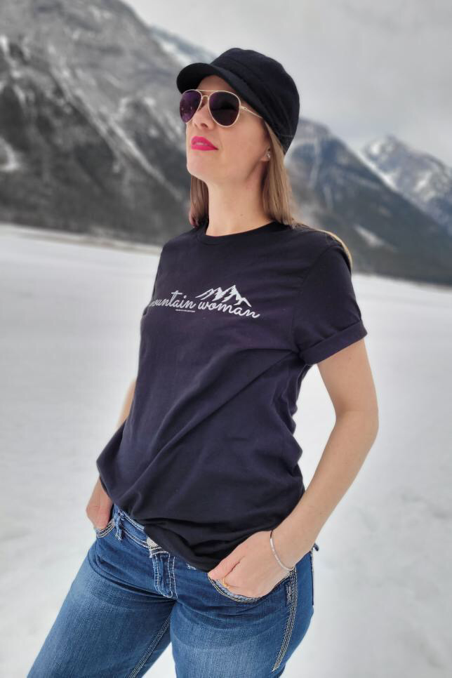 Woman wearing a black tshirt that says mountain woman standing outside with mountains in the background