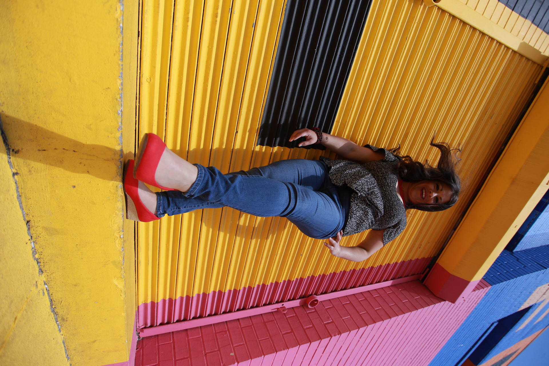 Wendie Veloz standing on a yellow curb with a colorful wall behind her wearing jeans and red shoes