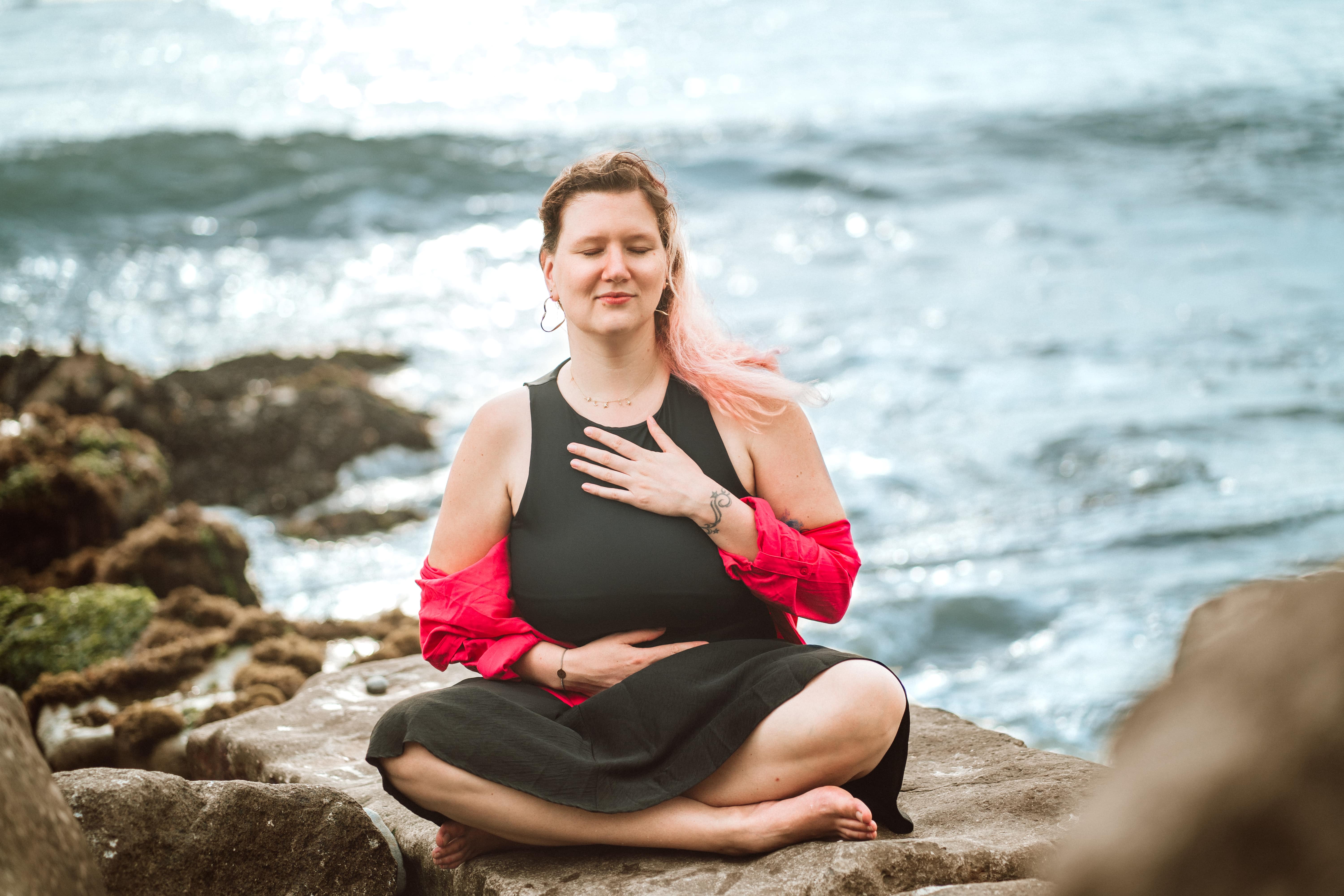 Foto von Svenja. Sie sitzt im Schneidersitz auf einem Felsen, das Meer im Hintergrund. Sie hat die Augen geschlossen und eine Hand auf ihrem Bauch, eine Hand auf ihrem Brustkorb, während sie eine Atemübung macht.