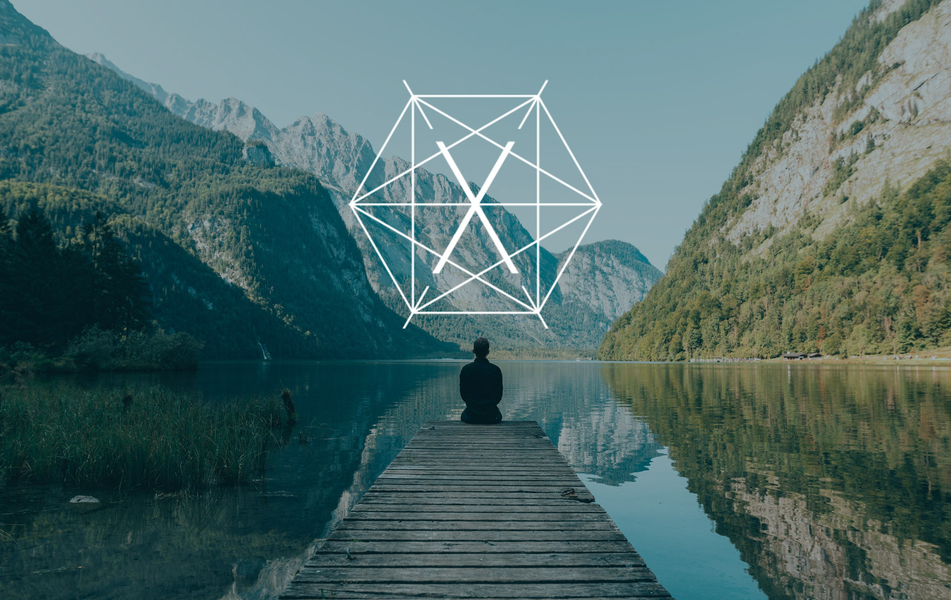 A man sits on a dock facing mountains and a still lake. His back is facing the camera