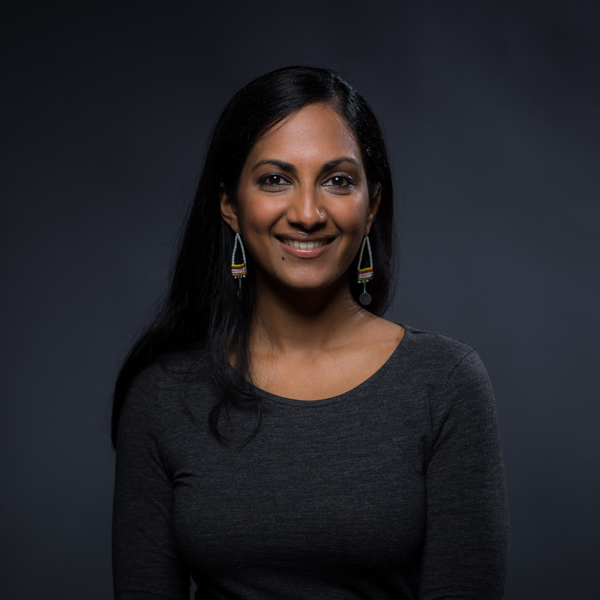 Young, smiling Indian American woman will long black hair, and long triangle-shaped earrings, and a dark gray cotton, long sleeved shirt. Background is dark gray. 