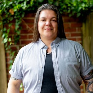 Genderqueer individual with long dark hair, wearing short sleeve button up shirt, colorful tattoos on arms. looking directly at the camera. They are outside with ivy covered wall in background. 