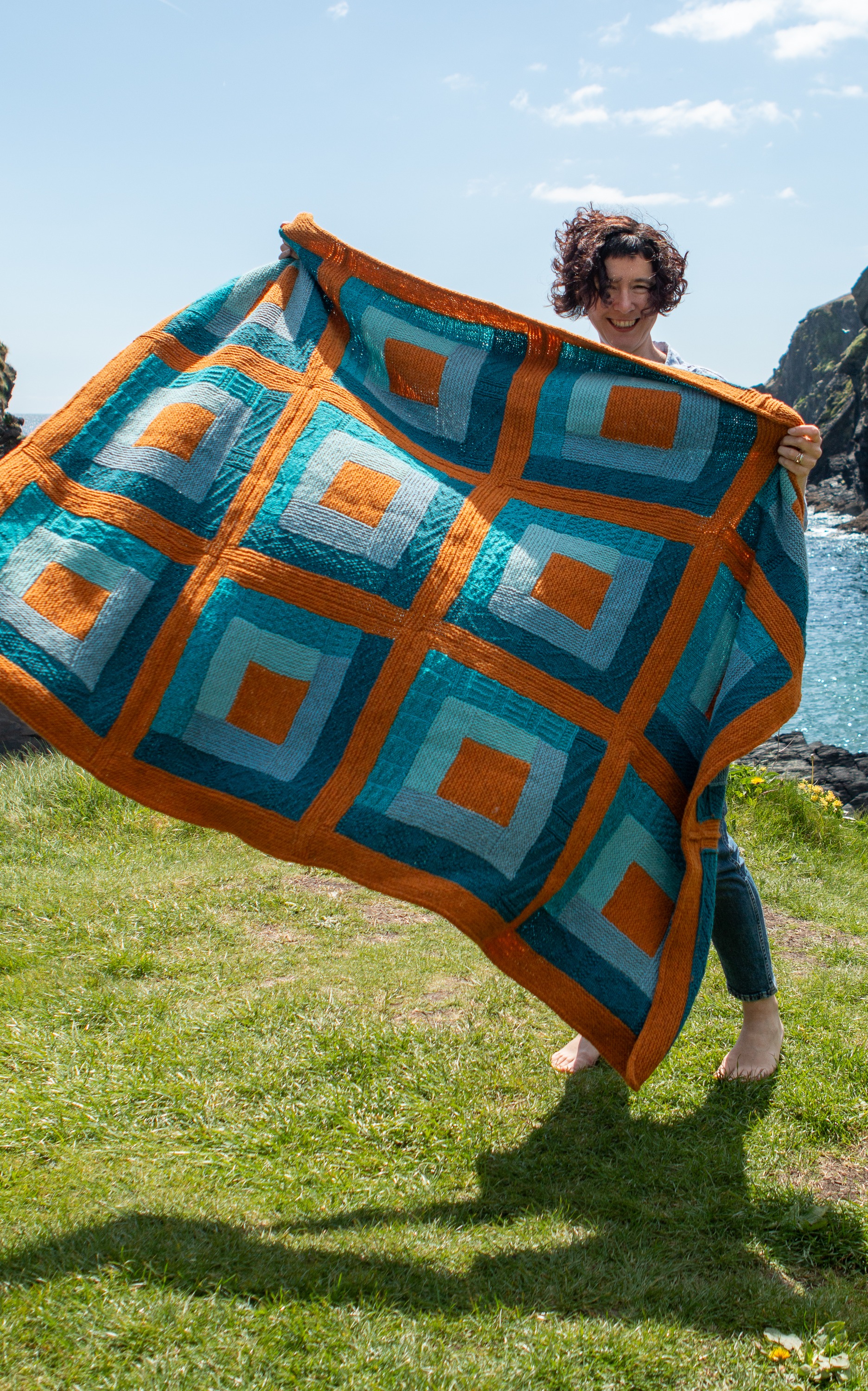 Woman holding a colourful log cabin style blanket up and smiling. 