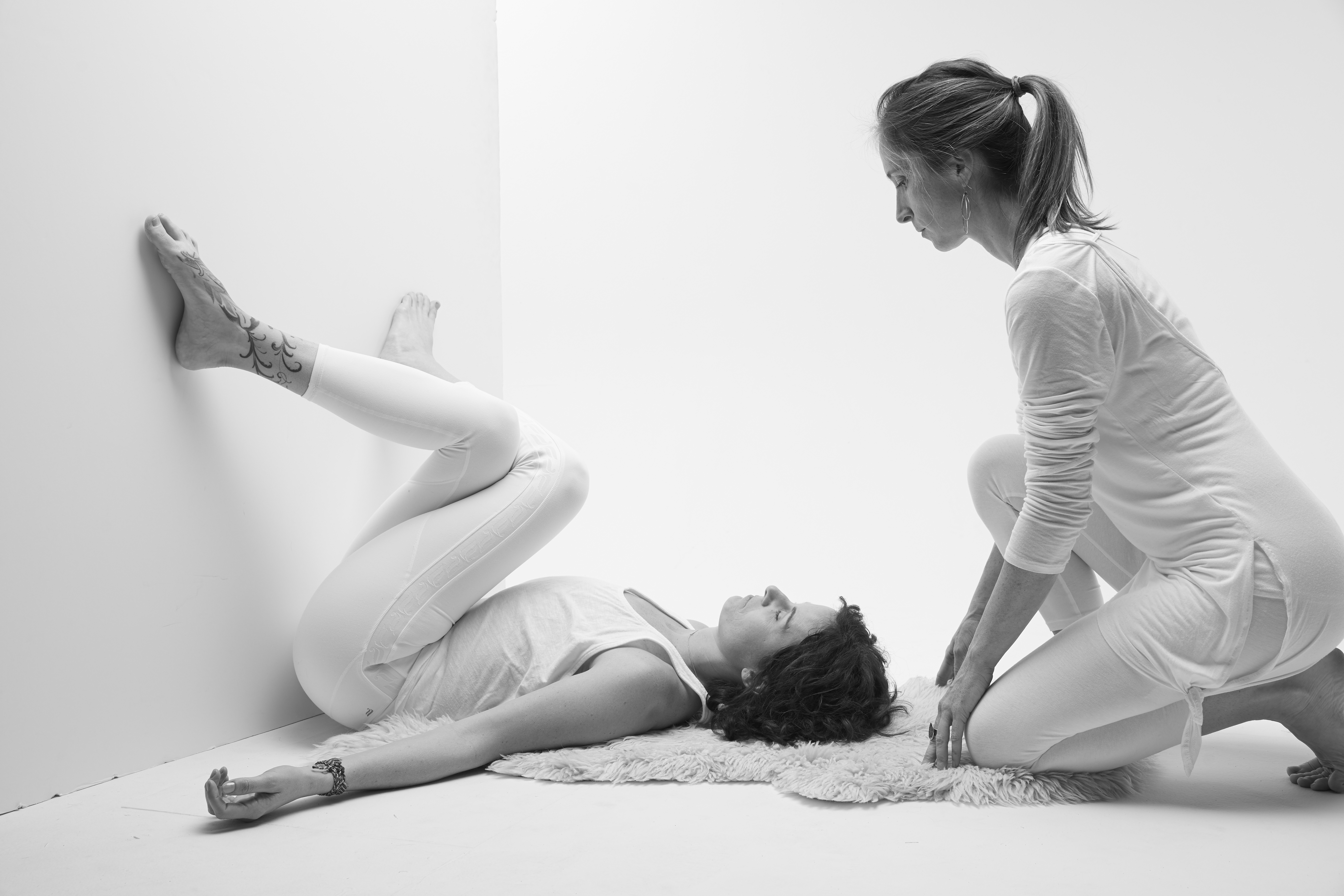 black and white image of yoga teacher Ivy Hughes demonstrating shoelace pose at the wall with Yoga teacher Kari Kwinn supporting
