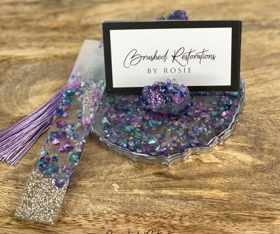 A handmade resin bookmark with delicate amethyst-toned tassels lies beside an ornate resin coaster infused with vibrant hues of purple and turquoise. The coaster features a sparkling, crystallized design, exuding a luxurious charm. A business card with the elegant script reads 