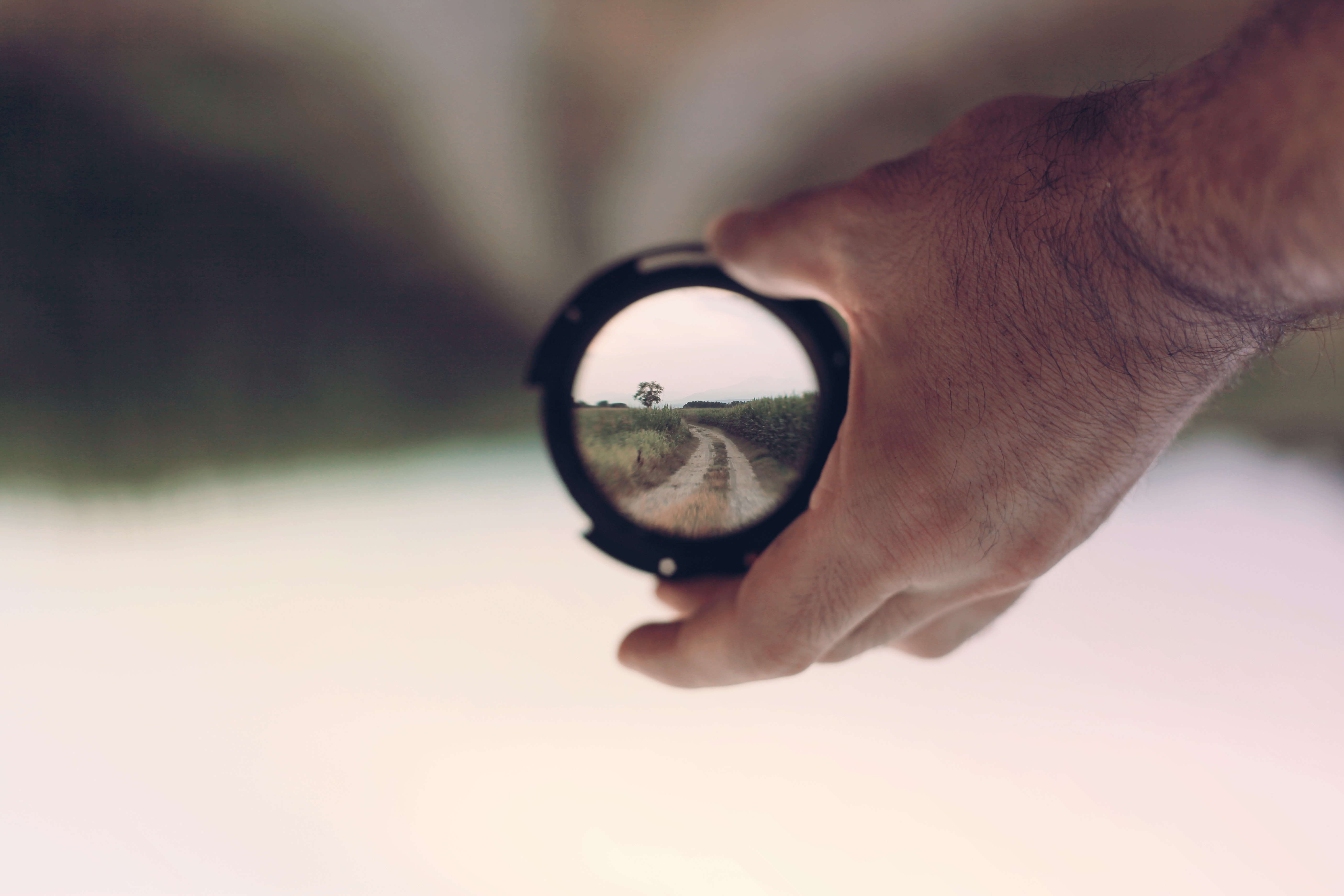 hand holding lens bringing blurry image into clear image of dirt road lined by tall greenery and a tree in distance