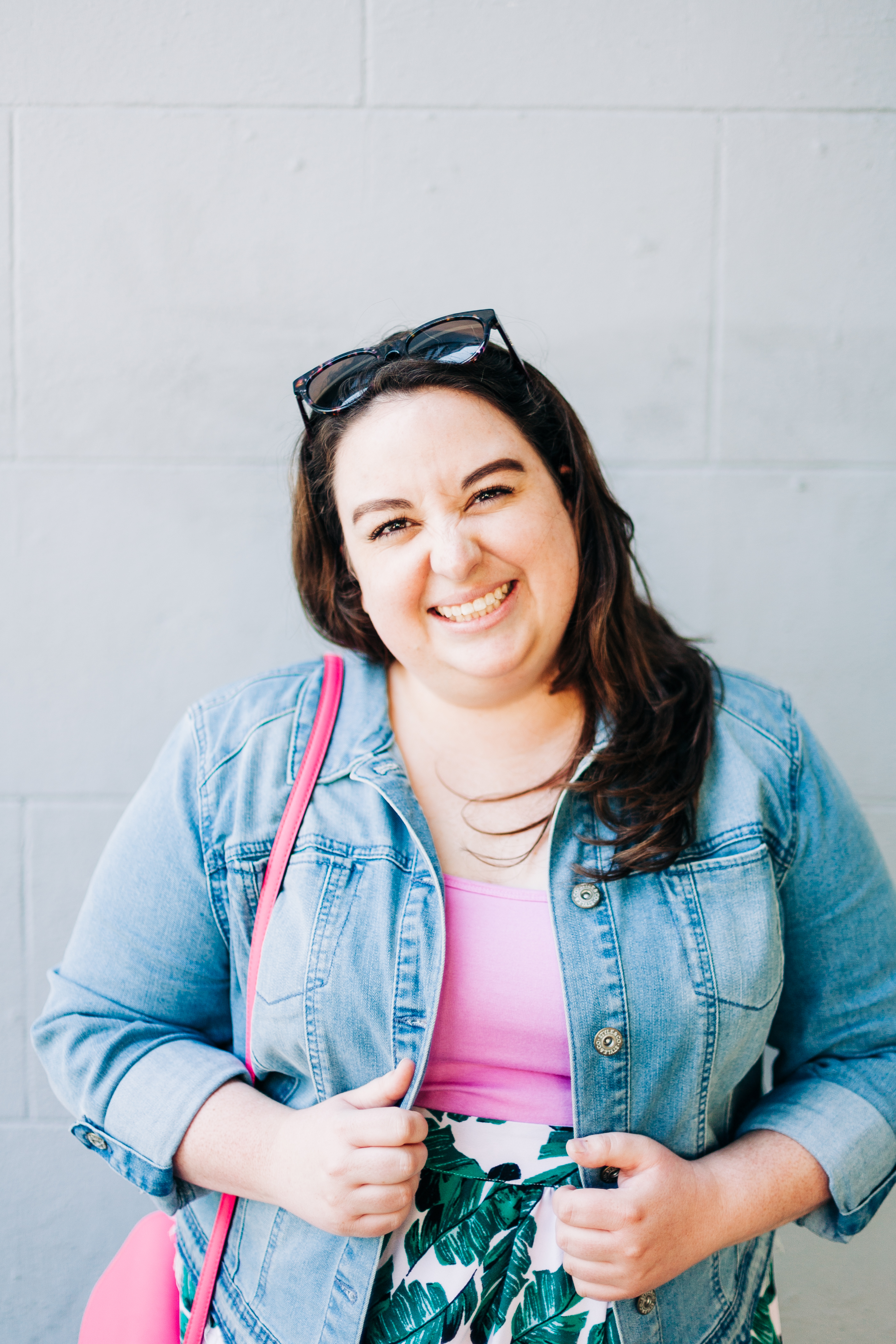 Kristen Achziger is standing in front of a gray brick wall. She is wearing a lavender shirt and a denim jacket and is smiling at the camera.