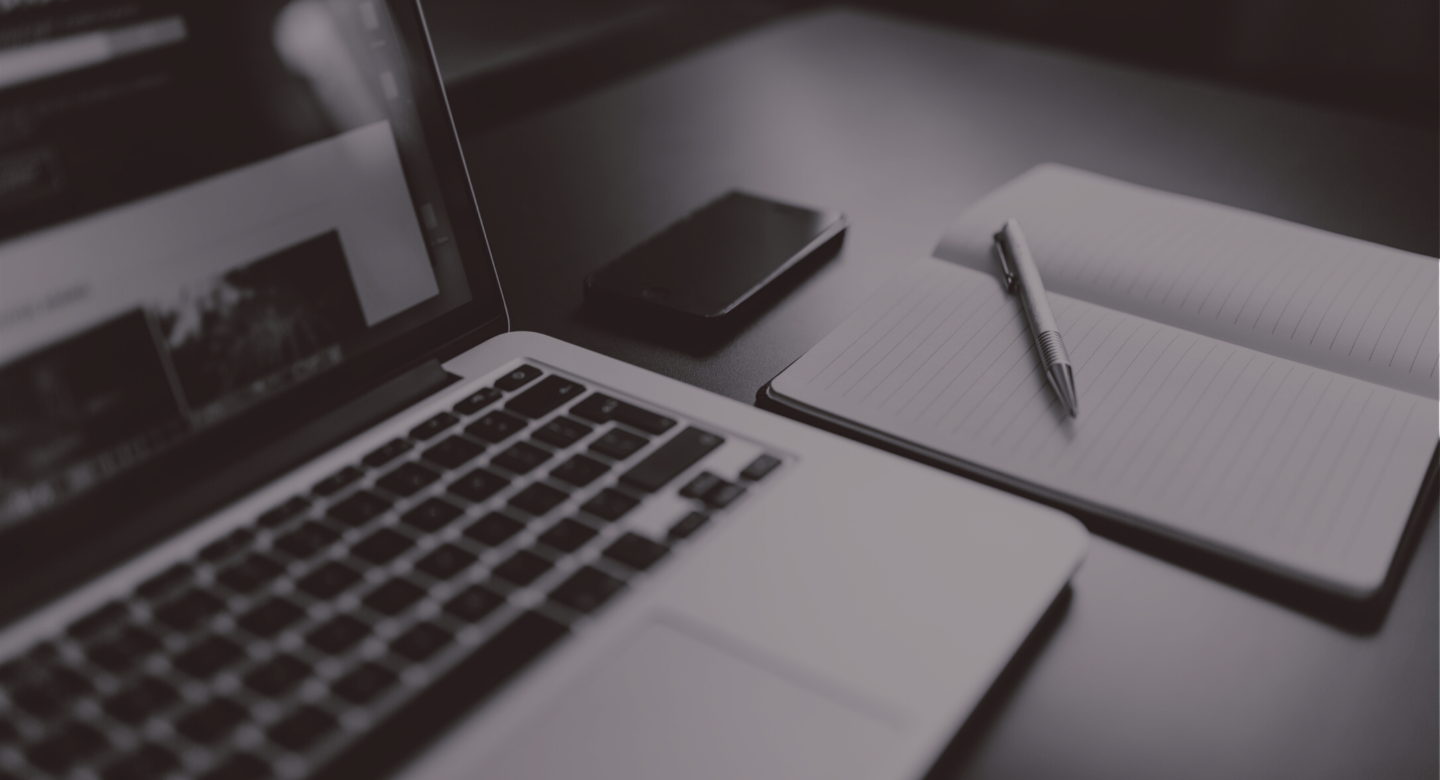 Laptop on desk next to mobile phone and notebook with pen.