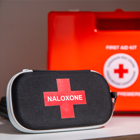 Une trousse de naloxone est posée sur une tablette devant une trousse de premiers soins de la Croix-Rouge.