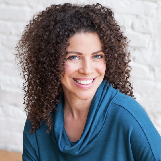 Woman with long, dark brown curly hair smiling at the camera wearing a teal loose collared turtleneck sweater with white brick wall in the background.