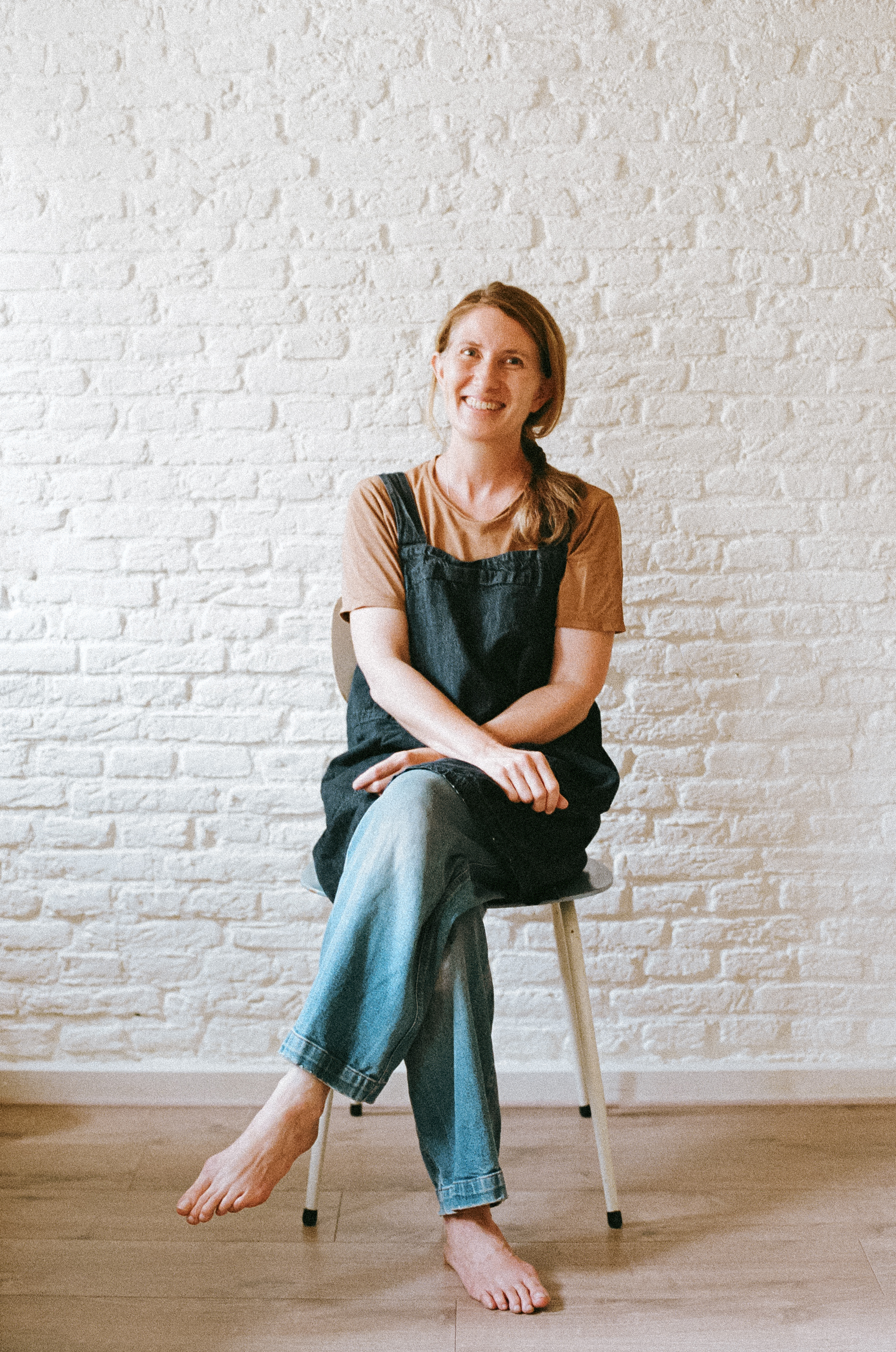 Picture of Simone sitting on a chair with legs crossed. She is wearing a brown tshirt, jeans and a blue apron