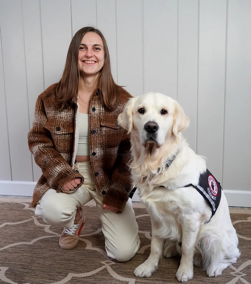 Light-skinned woman in brown jacket and creme pants kneels next to a light-colored golden retriever in a service dog vest
