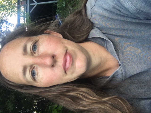 Rachel&#39;s headshot with her long brown hair loose, wearing a charcoal grey sweatshirt, sitting in front of greenery.