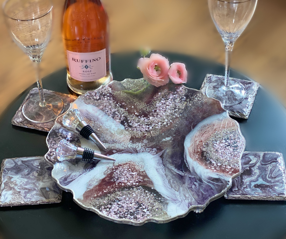 An exquisitely arranged resin art set serves as the centerpiece for a sophisticated wine tasting. A large, scalloped-edge resin tray, swirling with marbled whites, soft purples, and glitter accents, holds a bottle of Ruffino Rosé. Flanked by matching resin coasters adorned with bottle stoppers, the setup is completed with two delicate champagne flutes and tender pink roses, creating an ambiance of refined elegance.