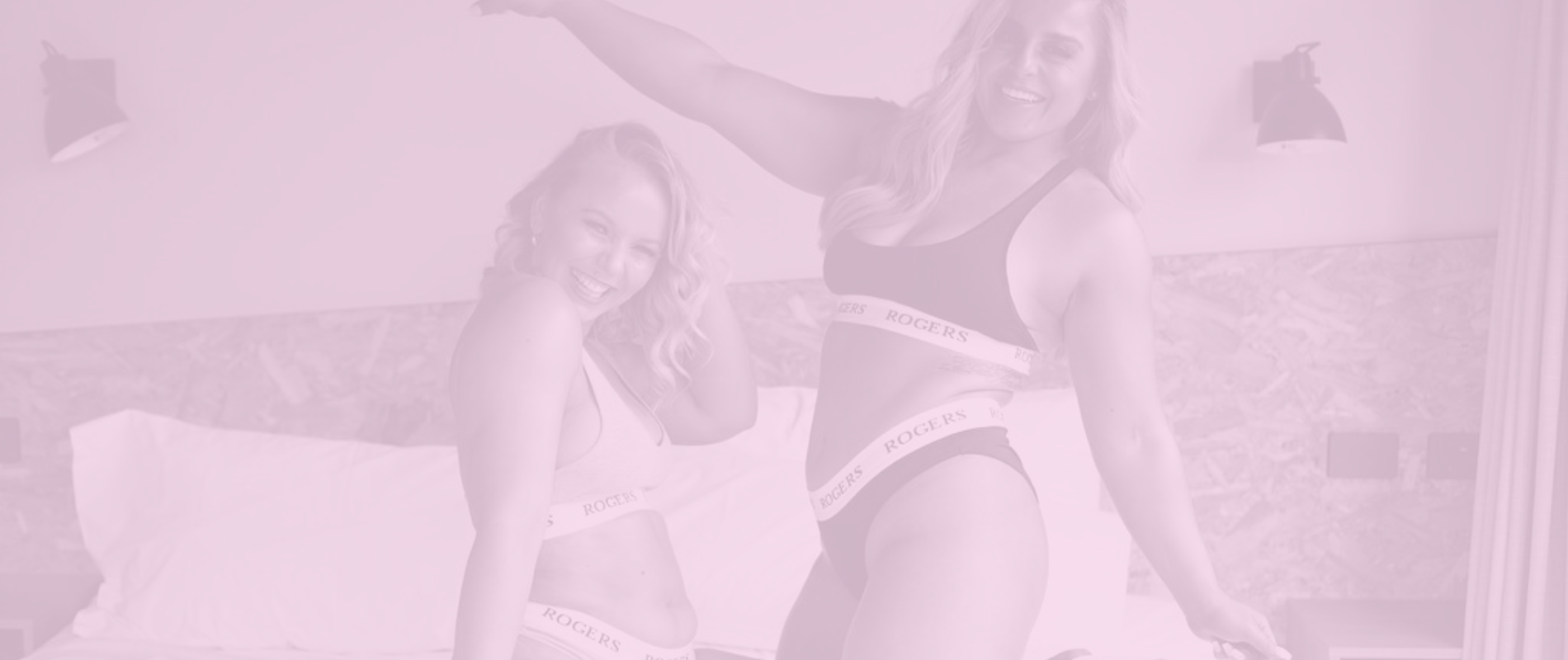 Two women on a bed in their underwear smiling and dancing, embracing their bodies