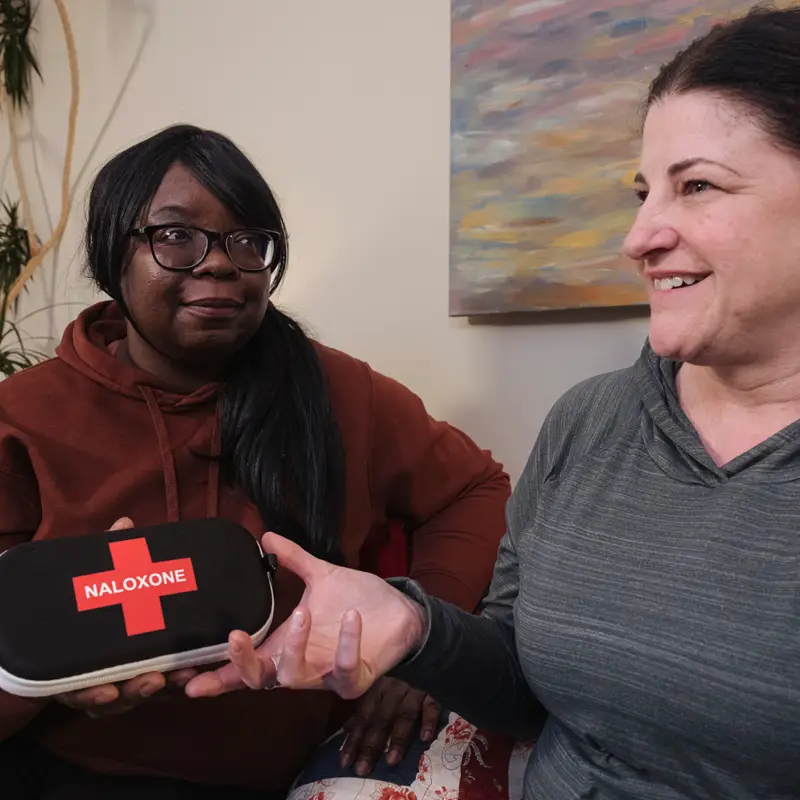 Two people having a conversation on a sofa while one shows a naloxone kit to the other. 