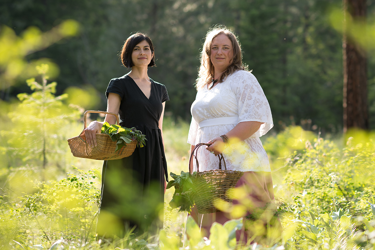 Emily Han and Rosalee de la Forêt standing among green plants and holding baskets of herbs