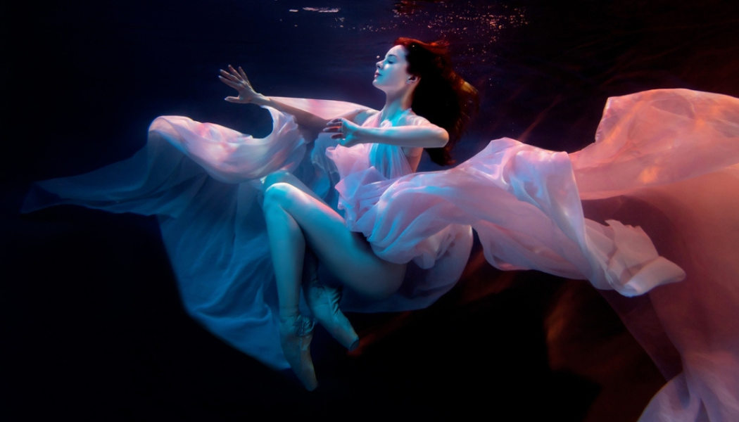 A woman in a flowing white dress submerged in a pool of water, with the dress flowing out behind her. 