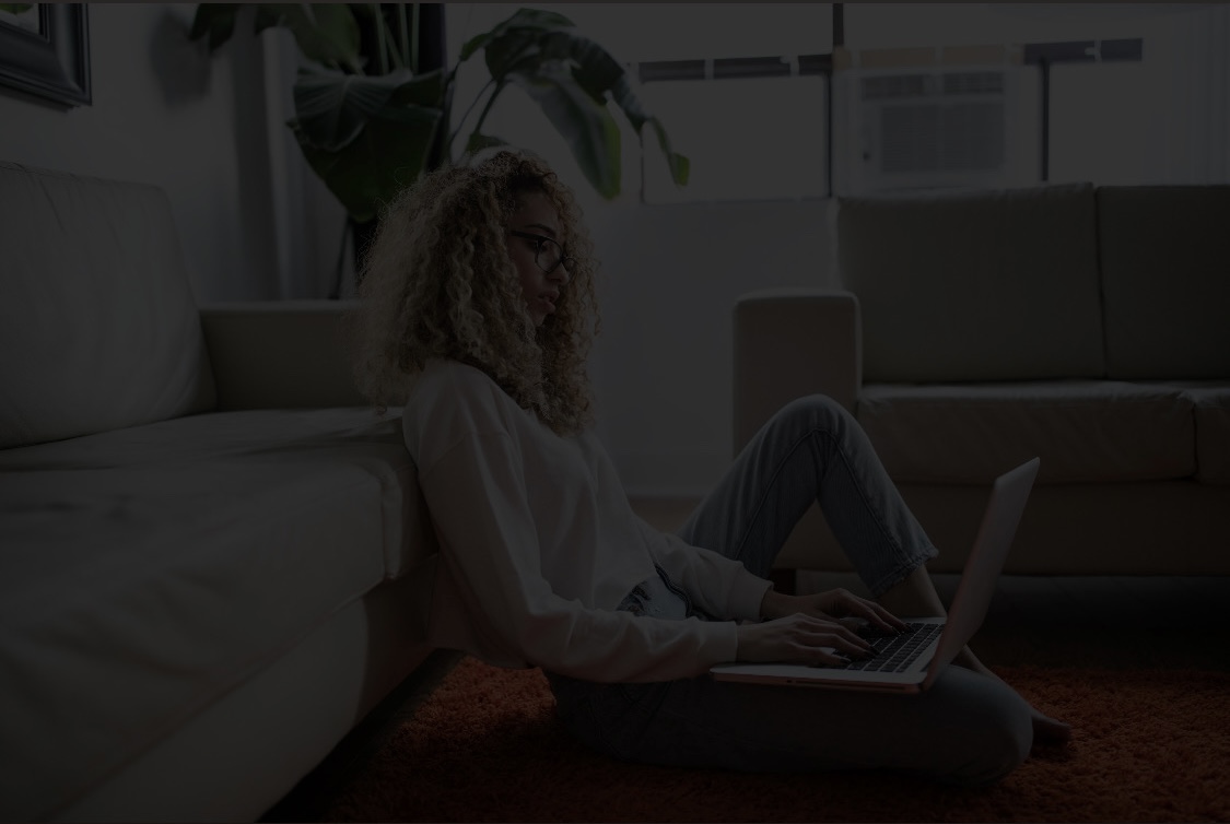 Woman with long curly hair sitting on floor in front of couch, typing on a laptop.