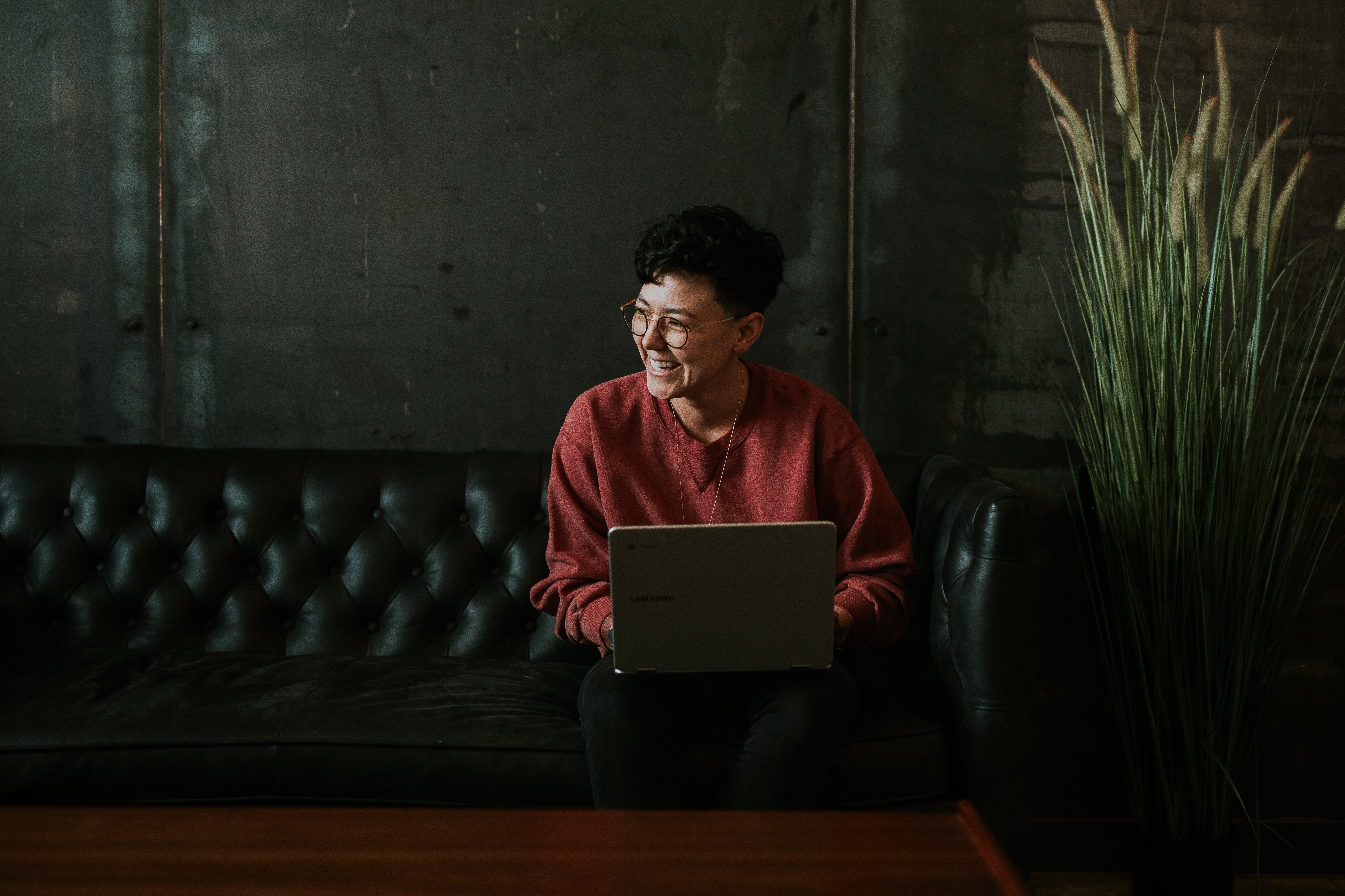 A woman with short hair and glasses smiles while sitting on a leather sofa with a laptop