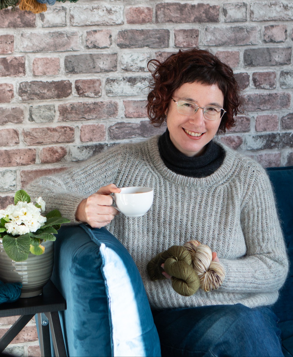 Carol Feller holding a cup of coffee and one of the yarn options while she is sat on a couch and smiling at the camera.