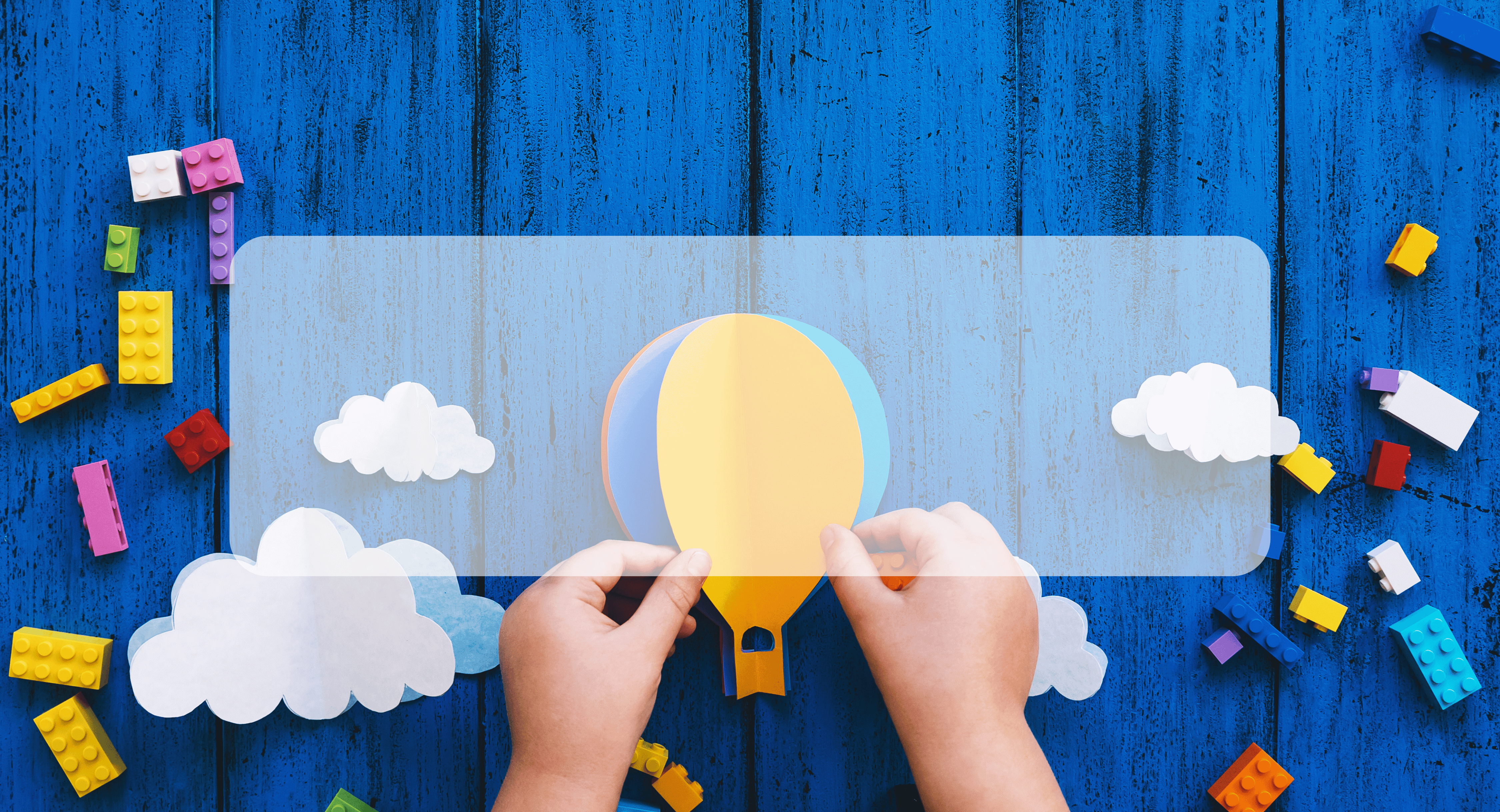 childs hands holding paper hot air balloon with paper clouds and legos on a blue background
